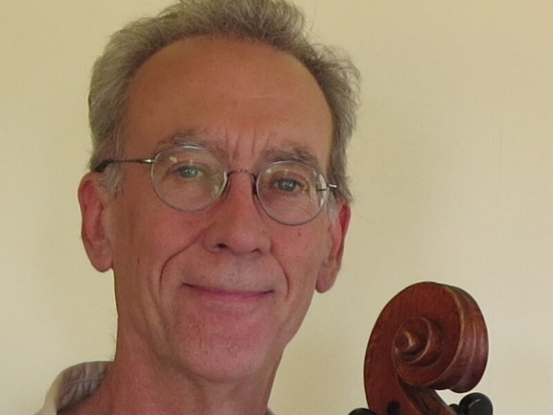 A man smiling at the camera with the scroll of a cello next to him