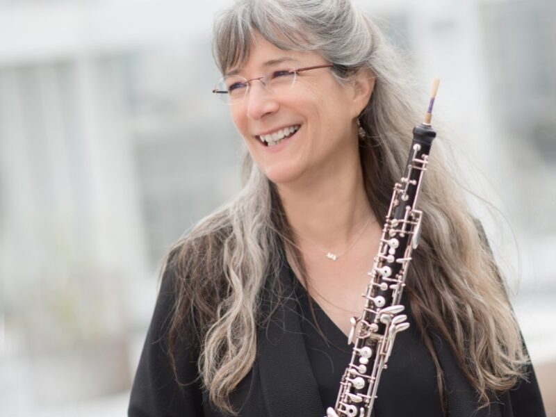 A woman with long grey hair smiling holding an oboe