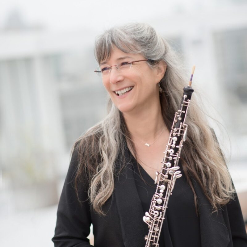 A woman with long grey hair smiling holding an oboe